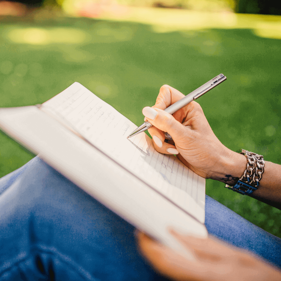 Woman writing in notebook outside with grass in the background.